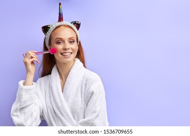 Attractive Caucasian Woman In Bathrobe Holding Makeup Bathrobe, Funny Hair Band On Head. Isolated Over Purple Background