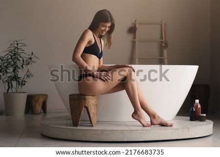Attractive Caucasian woman in bikini having morning spa procedures, touching her legs, enjoying smooth skin, sitting on low wooden stool near ceramic bathtub. Self-care, hygiene and relaxation