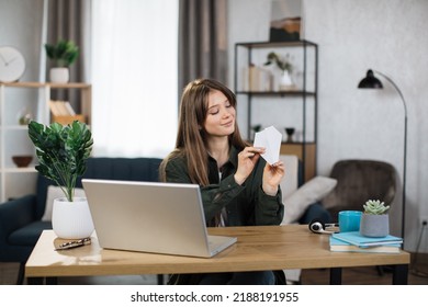 Attractive Caucasian Woman In Casual Outfit Sitting At Desk With Modern Laptop And Playing With Paper Plane. Female Boring At Work. Unproductive Concept.