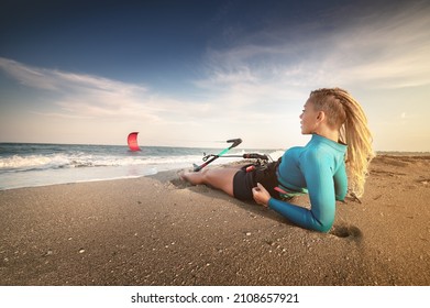 Attractive caucasian woman with dreadlocks on her head in a wetsuit lies on a sandy beach and holds her kite. Water sports. Kite surfer on vacation. Copy space - Powered by Shutterstock