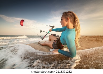 Attractive caucasian woman kitesurfer in a neoprene wetsuit and with dreadlocks on her head is resting lying on a sandy beach on the shore holding her kite in the wind - Powered by Shutterstock