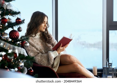 Attractive caucasian woman sitting near shiny christmas tree at home and spending free time for reading book. Cozy and relaxing atmosphere at modern apartment.