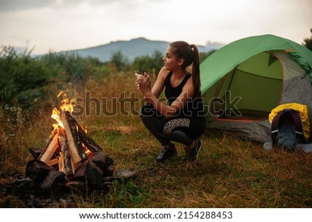 Attractive caucasian woman squatting near green tent and warming hands over bonfire. Active female alone on wild nature. Calmness and harmony concept.