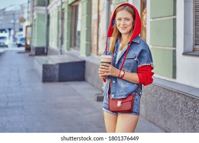 Attractive caucasian young cheerful woman about 25 years old in red hoodie and with long blond hair is drinking coffee from paper cup and smiling while walking in the center of Saint-Petersburg.