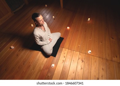 Attractive Caucasian young man meditates with closed eyes while sitting in lotus position in a dark room by candlelight.