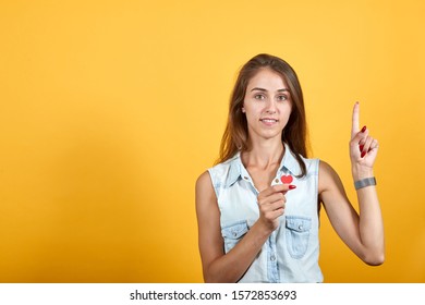 Attractive caucasian young woman in blue denim shirt pointing up, holding tiny red heart isolated on orange background in studio. People sincere emotions, lifestyle concept.