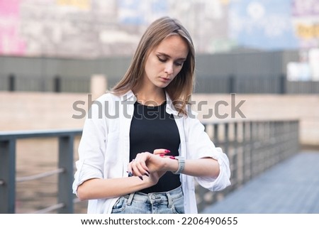 Attractive Caucasian young woman in casual clothing looking at a wristwatch while walking outdoors on a summer day. Concept of deadline and punctuality.