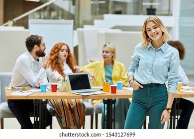 Attractive charming woman  looks at camera and engaging smile  keeps hand in pocket  poses against back ground working colleagues sitting at table in modern designers office
