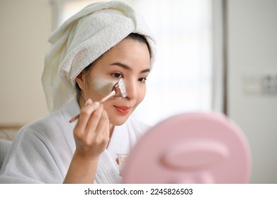 attractive and charming young Asian woman in bathrobe looking in the mirror, applying facial detoxifying clay mask, enjoying on her health care day at home. - Powered by Shutterstock