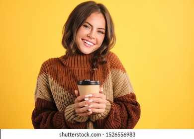 Attractive cheerful girl in cozy sweater with coffee to go happily looking in camera over yellow background