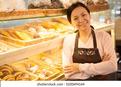 Attractive cheerful mature Asian female baker wearing an apron smiling joyfully to the camera posing at her bakery store copyspace small business owner entrepreneur cafe consumerism salesperson job. - Powered by Shutterstock