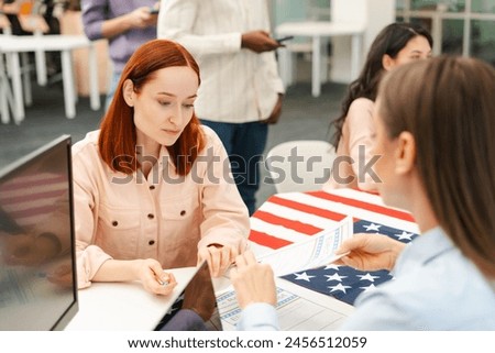 Attractive, cheerful woman with red hair talking to worker at polling station, sitting at registration table, writing down ballot paper. Concept of US presidential election