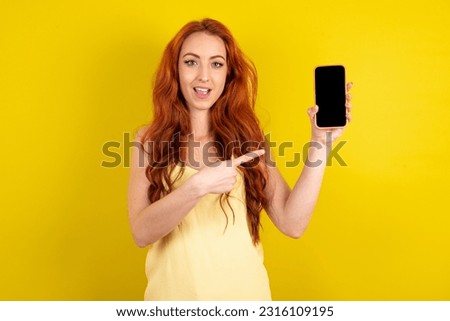 Attractive cheerful young beautiful red haired woman wearing yellow shirt over yellow studio background  holding in hands cell showing black screen