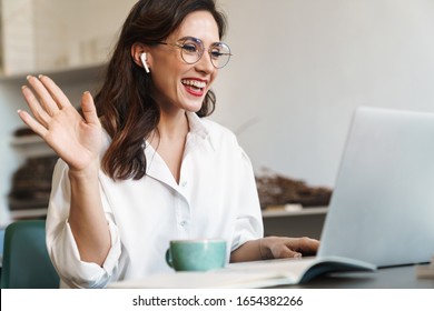 Attractive Cheerful Young Brunette Businesswoman Sitting At The Cafe Table With Laptop Computer Indoors, Wearing Wireless Earphones, Audio Call, Waving Hand