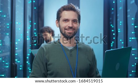 Attractive cheerful young server specialist using laptop coworking in data center providing webhosting with colleague staying at computer security.