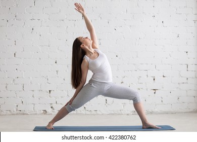 Attractive Cheerful Young Woman Working Out Indoors. Beautiful Model Doing Exercises On Blue Mat In Room With White Walls. Peaceful Or Reverse Warrior Pose, Viparita Virabhadrasana. Full Length