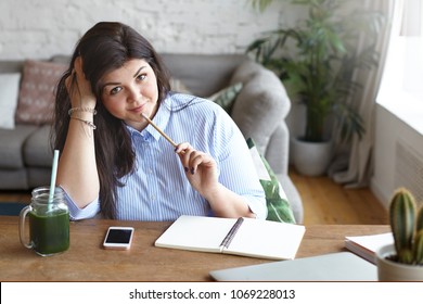 Attractive chubby student girl in blue shirt doing homework at desk, holding pencil at her chin, having pensive look while writing essay with green smoothie, diary and mobile phone in front of her