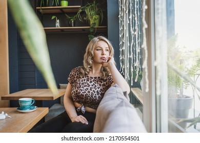 Attractive Chubby Young Blonde Woman In A Leopard Blouse Drinks Coffee From A Turquoise Cup In A Cafe Sits Near A Panoramic Window. Plus Size Girl With A Cup Have Fun 