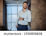 Attractive confident businessman in an elegant suit and a modern watch on his wrist. He is leaning on the wall with his art with wristwatch in an indoor business space. Seductive look and a smile