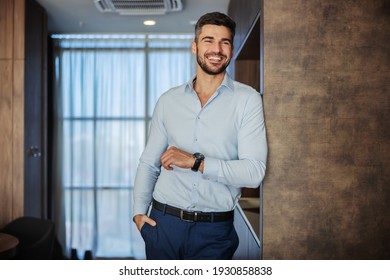 Attractive confident businessman in an elegant suit and a modern watch on his wrist. He is leaning on the wall with his art with wristwatch in an indoor business space. Seductive look and a smile