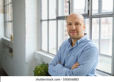 Attractive confident middle-aged man leaning against a windowsill in a relaxed position with folded arms staring thoughtfully at the camera, with copy space - Powered by Shutterstock