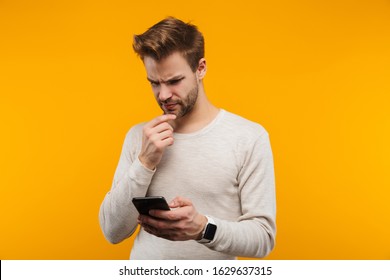 Attractive Confused Young Man Wearing Pullower Standing Isolated Over Yellow Background, Using Mobile Phone