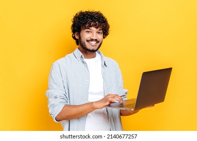 Attractive contented indian or arabian curly haired guy dressed in casual wear, holding an open laptop in his hand, looks friendly smiling at the camera, while standing on isolated orange background - Powered by Shutterstock