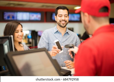 Attractive Couple Buying Tickets At The Movie Theater And Using A Smartphone To Show A Barcode To An Employee