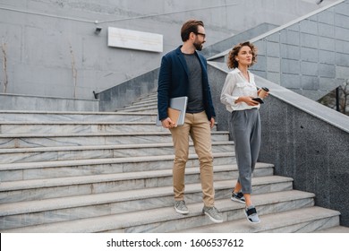 attractive couple of man and woman going on stairs in urban city center in smart casual business style, talking, working together, smiling, stylish freelance people, holding laptop, discussing
