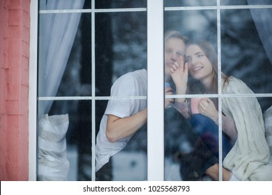 Attractive couple stay at the window. A girl is trying to show a boy falling snowflakes.