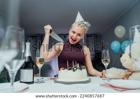 Attractive crazy laughing birthday woman with bread-knife planning to cut her birthday cake while celebrating birthday at home alone. Selective focus.