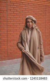 Attractive curly blonde woman dressed beret, coat, scarf and sweatshirt. Total beige or brown look. Background brick wall. Street fashion for spring, winter or fall season. Minimalistic look. - Shutterstock ID 1245118483