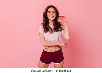 Attractive curly dark-haired woman in t-shirt and shorts is smiling on pink background