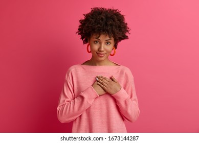 Attractive curly haired beautiful woman feels thankful, presses palms to chest, looks with gratitude, hears heartwarning supporting words has confident calm expression wears rosy jumper, being touched