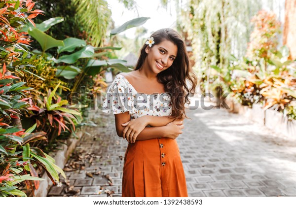 オレンジのスカートをはいた魅力的な巻き毛女性と 笑顔でカメラを見る白い上部 植物園で髪のポーズに花を持つ女の子 の写真素材 今すぐ編集