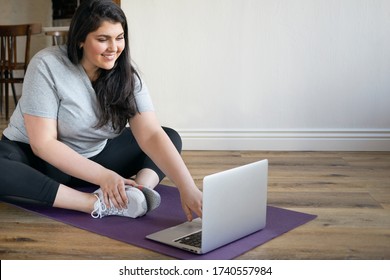 Attractive Cute Girl With Curvy Body Sitting On Mat Exercising To Lose Weight Using Laptop, Doing Distant Training Via Video Conference Chat Under Supervision Of Professional Fitness Instructor