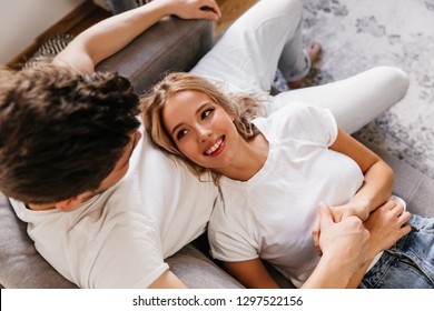 Attractive cute girl looks at her boyfriend with love in her eyes while laying on his legs. Sweet photo from above with couple holding hands at apartments.