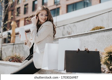 Attraktives süßes Mädchen mit den Einkaufstaschen und warmen Drinks in der Hand. Moderne Stadtmegapolis auf Hintergrund. Lady bestellen und online einkaufen. Schöne Kaukasierin