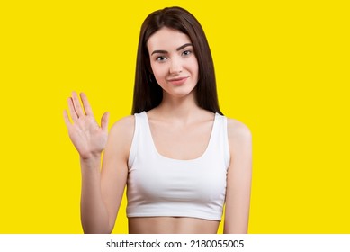 Attractive Cute Girl Waving Hello In A White Top On A Yellow Background. Shows Palm