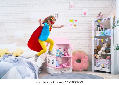 Attractive Cute Little Girl Jumping From Bed To Fly, She Plays Superhero With Cloak And Mask At Home In Kids Bedroom.concept Of Bravery And Courage In Face Of Small Child.feeling Of Freedom In Flight