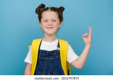 Attractive cute smiling schoolgirl showing okay gesture, wears yellow backpack. Close up portrait happy child accepting idea, posing isolated on blue studio background. Agreement, approval concept. - Shutterstock ID 2015797874