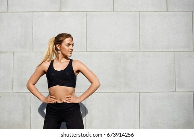 Attractive cute young female fitness instructor with ponytail dressed in black sportswear standing outdoors, keeping hands on her waist, making up her mind before workout class in the morning