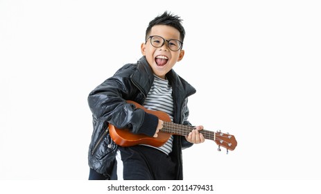 Attractive cutout portrait of smart young Asian boy wearing glasses, black jacket, and long pants happily dancing and enjoy acting as rock musician star by singing and playing small brown guitar