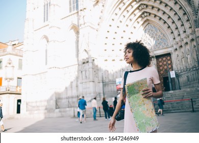 Attractive dark skinned traveler with paper map enjoying city sightseeing around ancient heritage of touristic urbanity, young female photographer exploring world during summer vacations for recreate