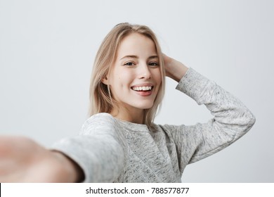 Attractive dark-eyed blonde female dressed casually having delightful look smiling broadly. Beautiful woman having cheerful expression while posing against gray background, stretching arm to camera.