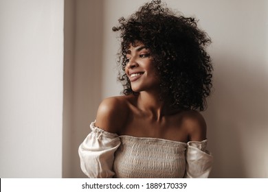 Attractive dark-skinned woman leans on white wall. Pretty curly brunette lady in stylish top smiles sincerely.