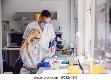 Attractive dedicated blond female lab assistant with face mask and rubber gloves doing research for the cure for corona virus. Next to her is her colleague standing and watching her.