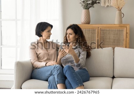 Attractive different age women, mature mom and young adult daughter lead trustworthy talk seated together on sofa, tell personal news, issues, receive support, asks advice, share life troubles at home