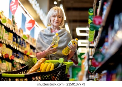 Attractive Elderly Woman Choosing Organic Oil At Supermarket, Female Pensioner Walking By Store With Trolley, Holding Two Bottles, Choosing Best Quality Food, Doing Grocery Alone