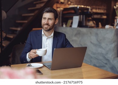 Attractive Elegant Businessman Holding Cup Of Coffee And Smiling At Laptop In Cafeteria. Portrait.